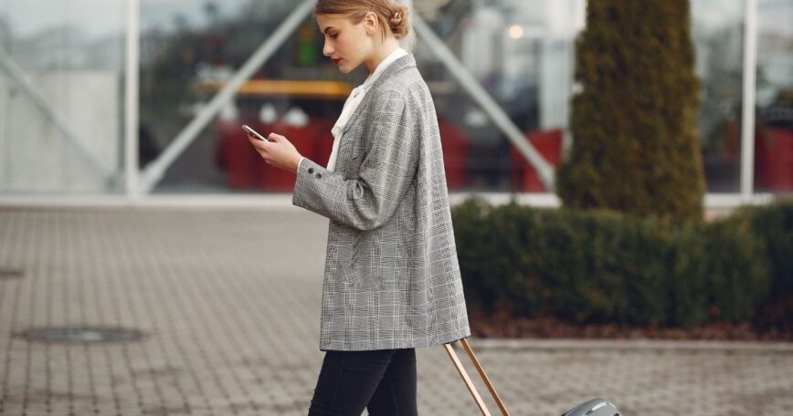 Traveler checking her phone while pulling a suitcase outside an airport